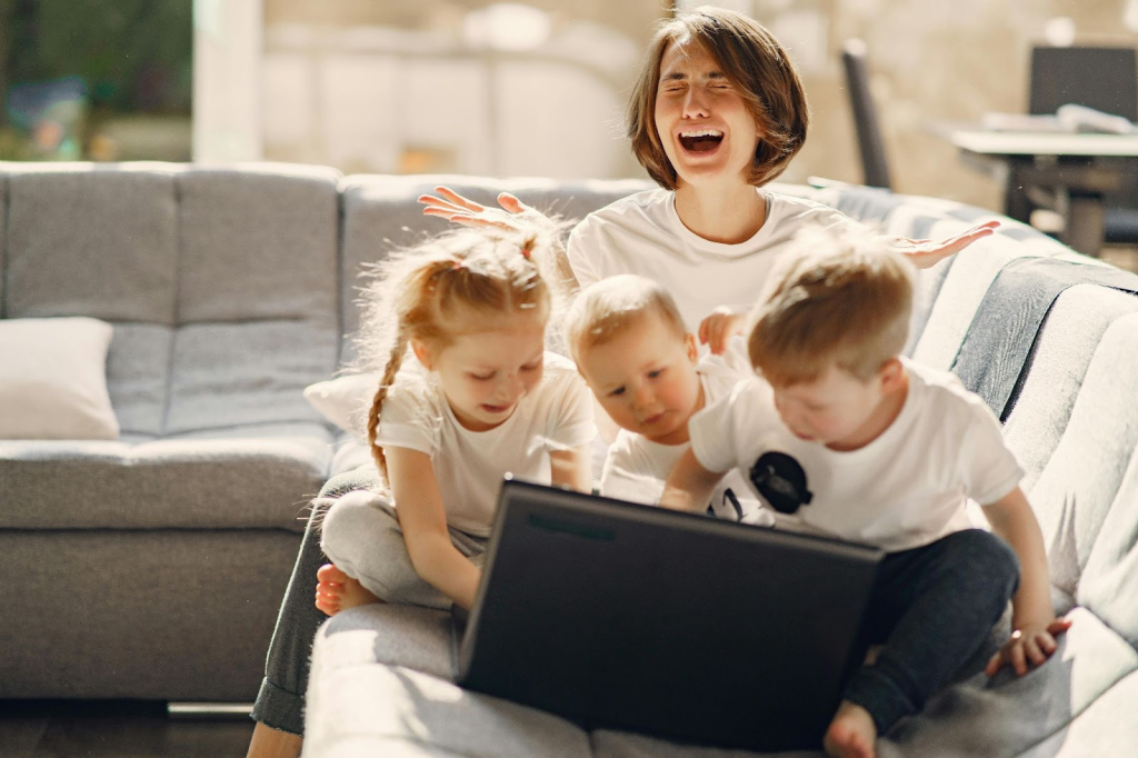 A post-Mormon mom on the couch crying with her hands in the air. Her three young children are on her lap all playing on her laptop her laptop. Illustrating how parents can have meltdowns and still parent from unconditional love.