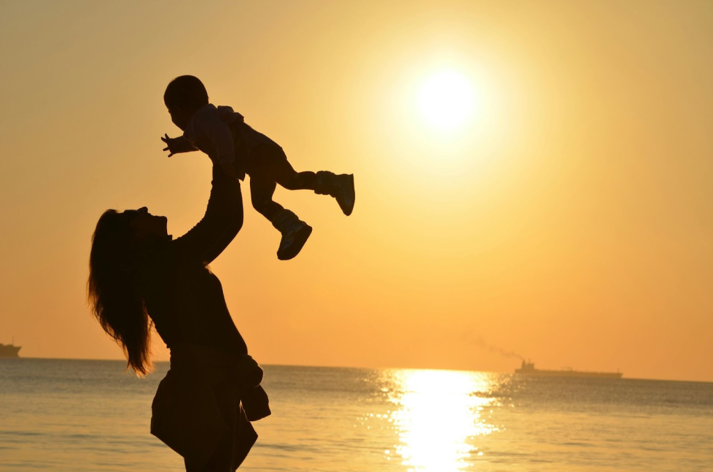 A mom holding her baby in the air with a beautiful ocean sunset behind her. Illustrating the fantasy that post-mormon parenting from unconditional love will be beautiful and easy.