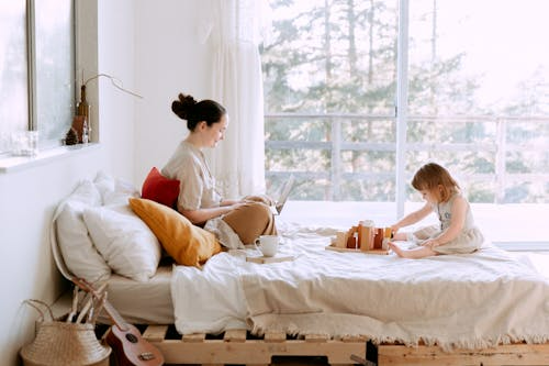 A mom working on her laptop on her bed with her little girl playing nearby. Illustrating connection in post-Mormon parenting