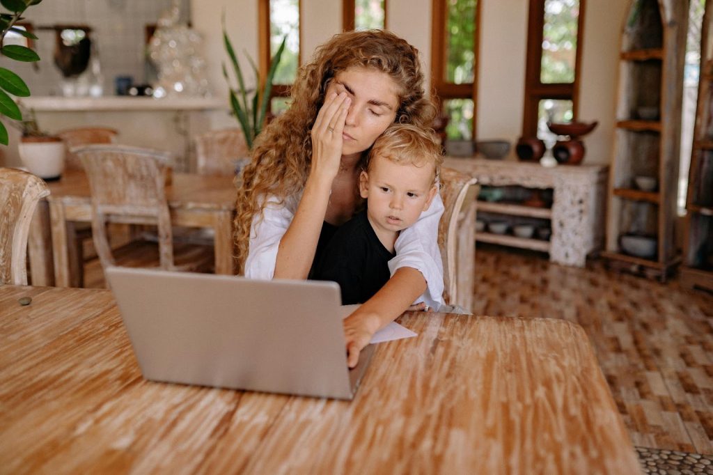 A mom sitting at the table rubbing her face in exhaustion with her son on her lap while she works on her laptop. Illustrating the exhaustion that post-Mormon parents can feel even when trying to parent from unconditional love.