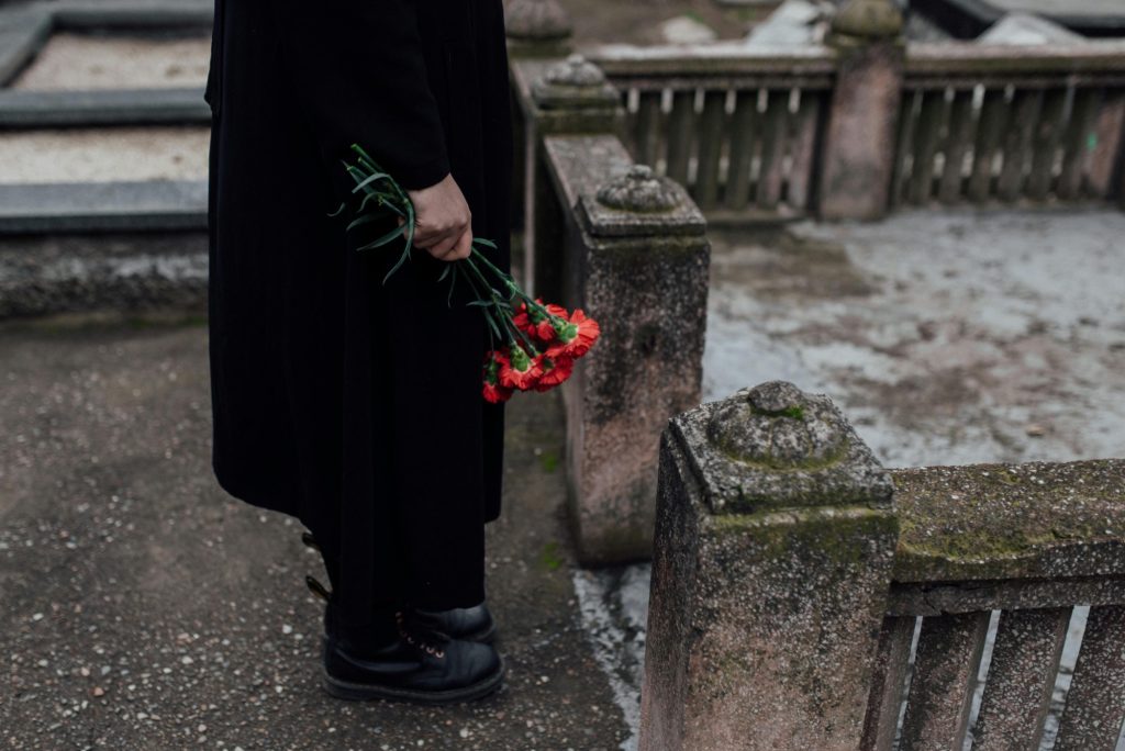 The bottom half of a person standing in front of a grave in a churchyard on a cold, dreary day. They are dressed in black and carry a bundle of red carnations. Illustrating the fear and bleakness of death.