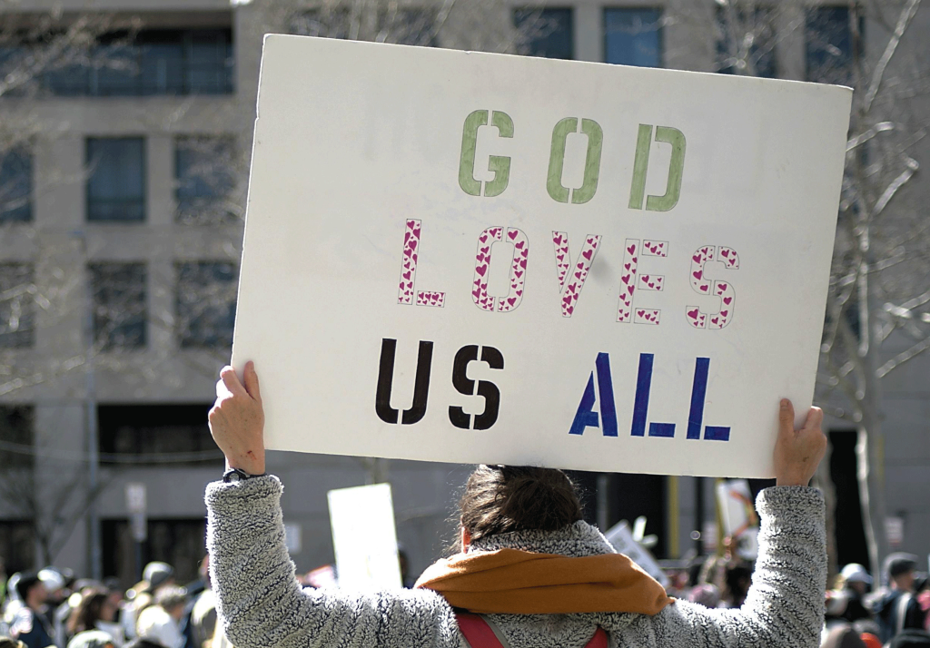 A woman at a protest holding a poster reading "God Loves Us All". Illustrates the love for humanity that flooded me after I left the LDS church.