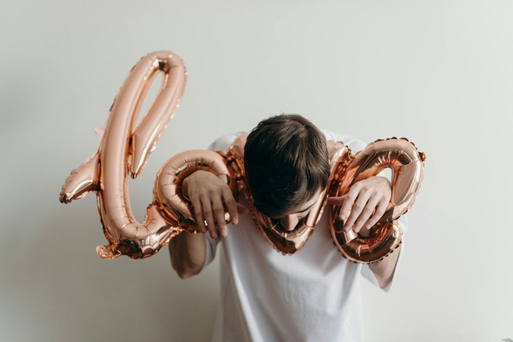 A balloon in the shape of the word Love. A man has his hands through the holes of the O and the E and his head in the V. It looks like he's in the stocks. Illustrating the restrictive and controlling conditional 'love' found in the LDS church.