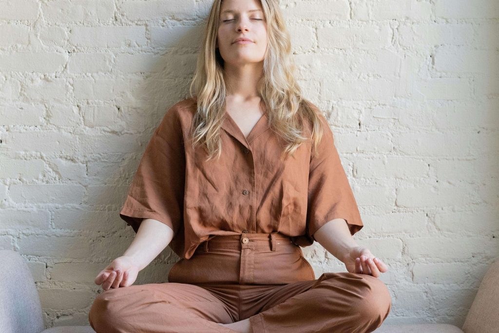 A woman sitting cross-legged against a wall with her hands resting on her knees with a peaceful smile. Illustrating how I found myself just sitting with God's love.